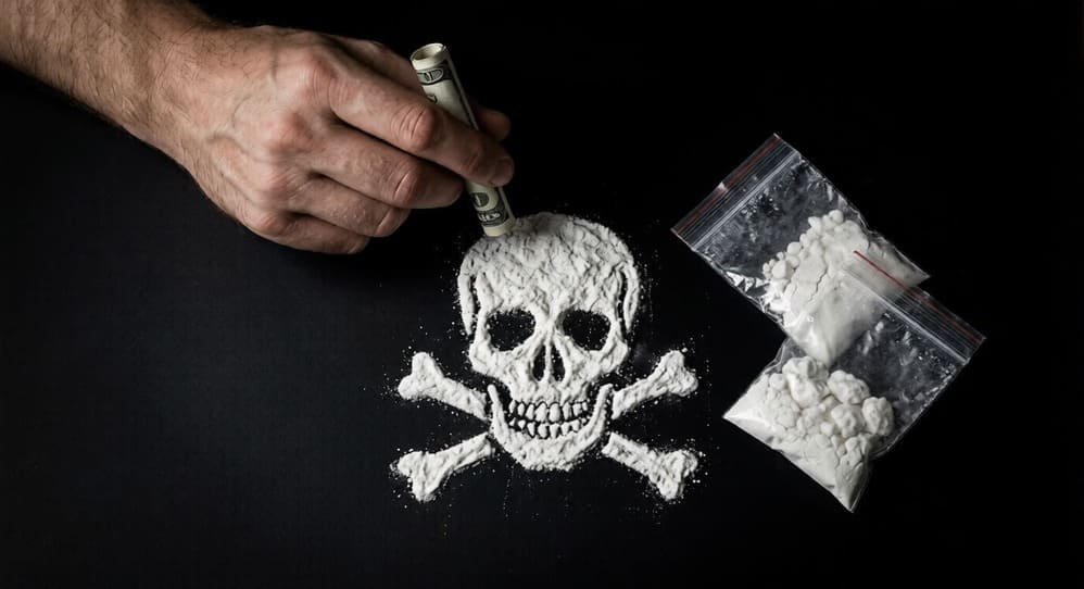 White powder shaped as a skull and crossbones next to drug bags and a hand holding money.